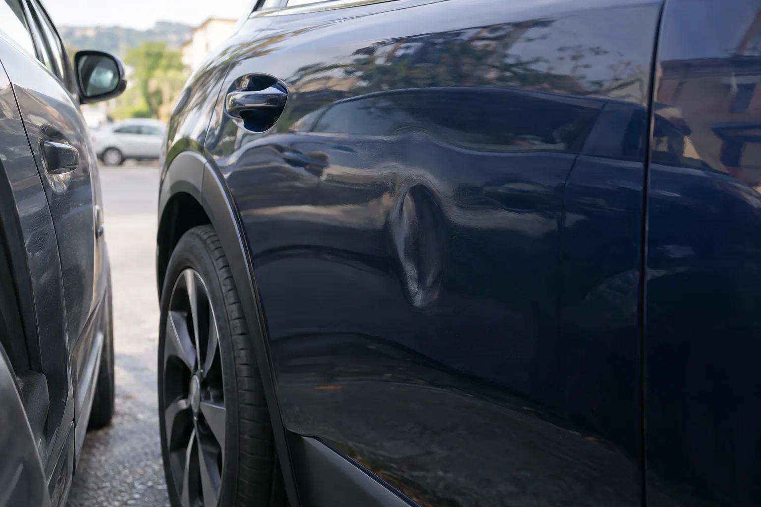 Gros plan sur une petite bosse sur la portière d'une voiture bleu nuit garée entre deux autres véhicules.